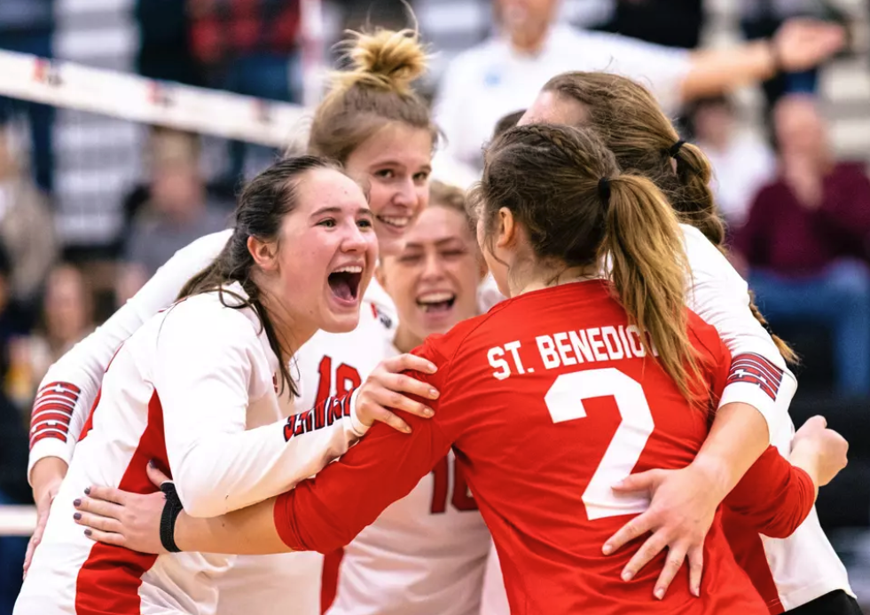 A group of female volleyball players wearing white and red jerseys celebrate together on the court. One player wearing a red jersey labeled "St. Benedict" is facing away from the camera. They appear joyful and energetic.